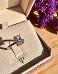 A gold ring with a radiant cut white diamond displayed inside an open blush pink velvet jewelry box on a wooden surface, with purple flowers in the background.