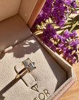 A gold ring with a radiant cut white diamond displayed inside a pink jewelry box, with purple flowers in the background on a wooden surface.
