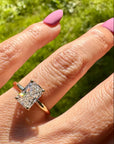 A gold ring with a radiant cut lab grown diamond worn on a person's ring finger, shown outdoors against a bright green grassy background.