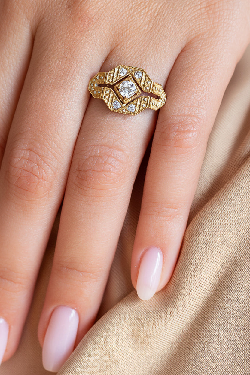 A close-up of a hand wearing an Art Deco style diamond ring. The ring features a central round diamond surrounded by geometric, beaded diamond bands in 14k yellow gold. 