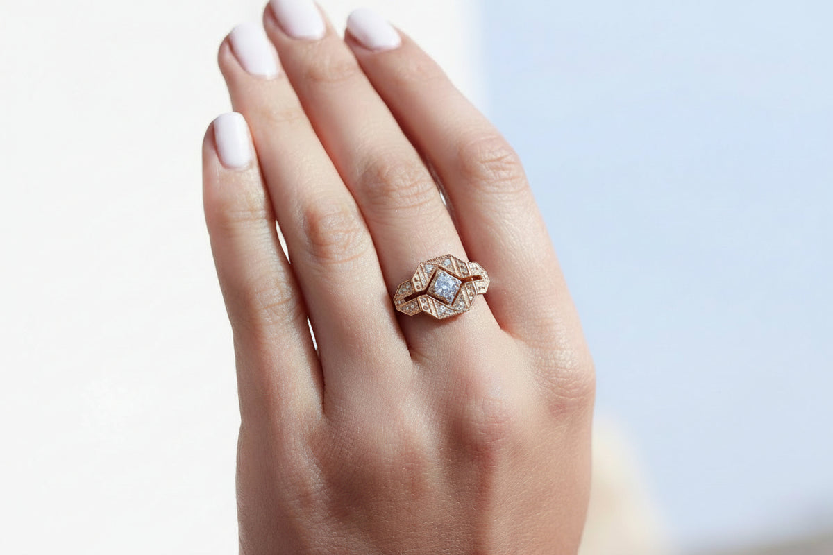 A 14k rose gold ring with a geometric, art deco style diamond-like pattern, set with one center brilliant cut white diamond and four smaller ones,  worn on a hand with pale pink manicured nails, set against a soft, out-of-focus light blue and white background.