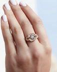 A 14k rose gold ring with a geometric, art deco style diamond-like pattern, set with one center brilliant cut white diamond and four smaller ones,  worn on a hand with pale pink manicured nails, set against a soft, out-of-focus light blue and white background.