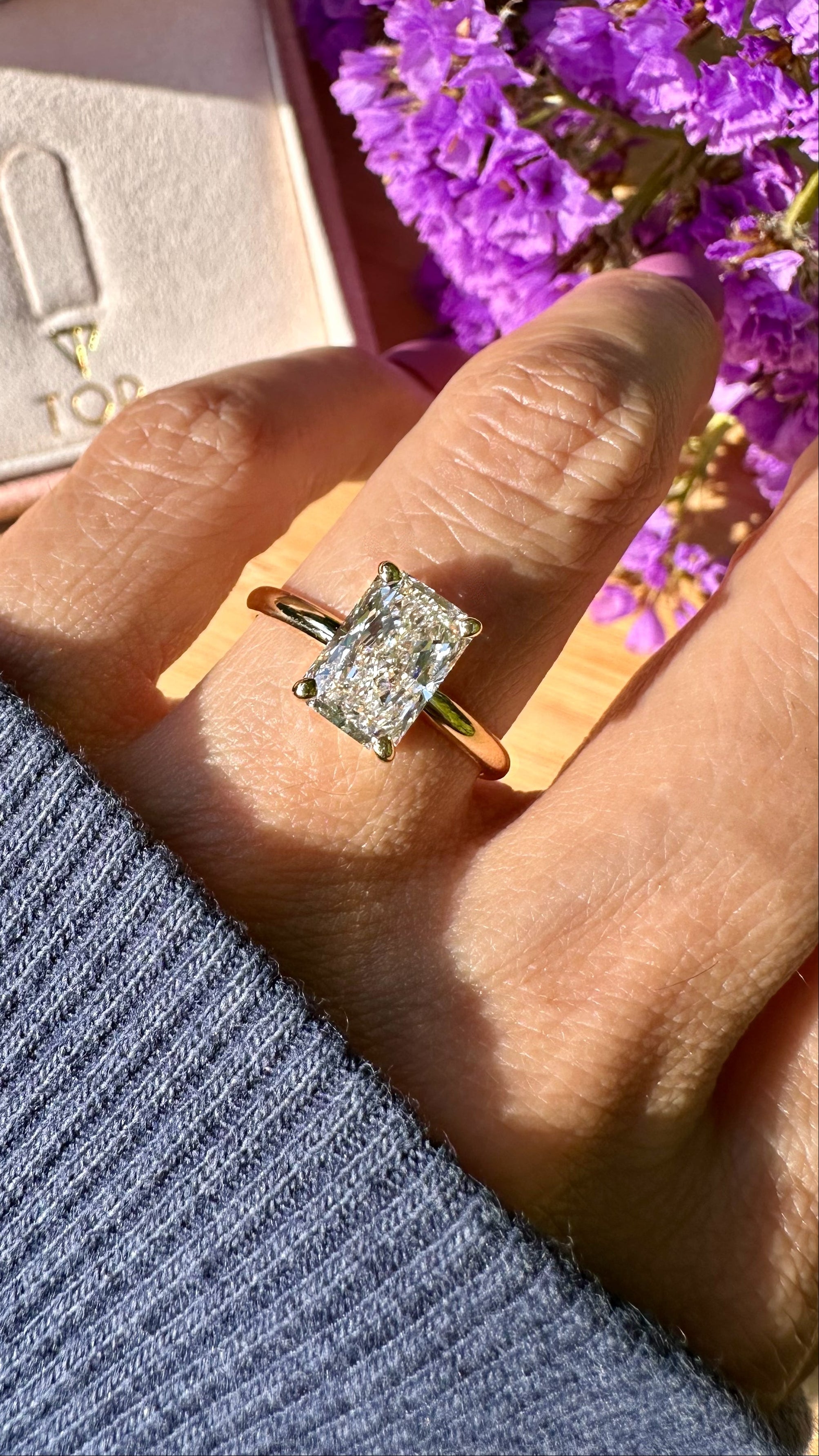A close-up lifestyle shot of a hand wearing a yellow gold ring with a radiant cut white diamond. The background features a pink jewelry box with a gold TOR logo and purple flowers.