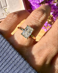 A close-up lifestyle shot of a hand wearing a yellow gold ring with a radiant cut white diamond. The background features a pink jewelry box with a gold TOR logo and purple flowers.