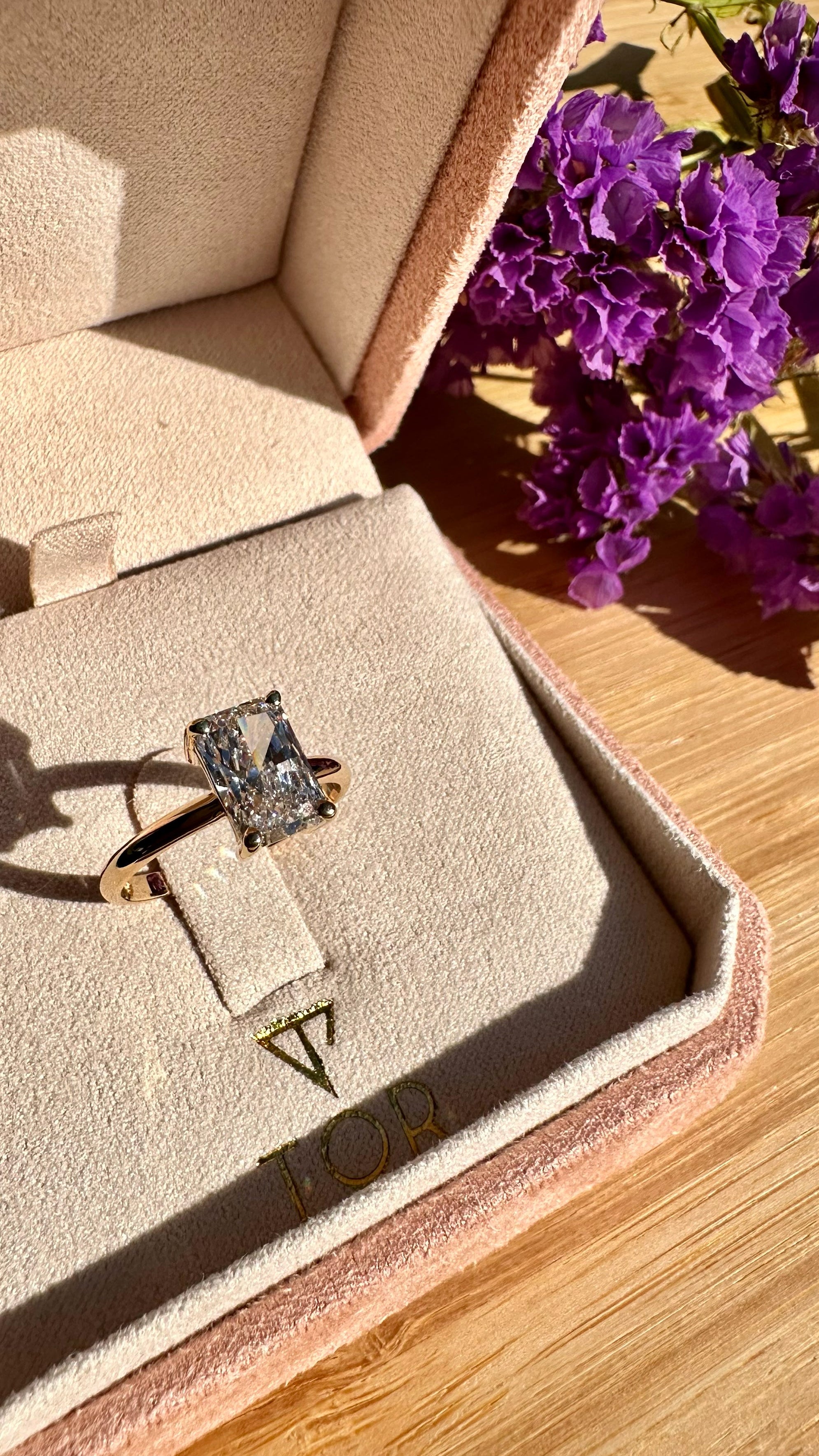 A gold ring with a radiant cut white diamond displayed inside an open blush pink velvet jewelry box on a wooden surface, with purple flowers in the background.