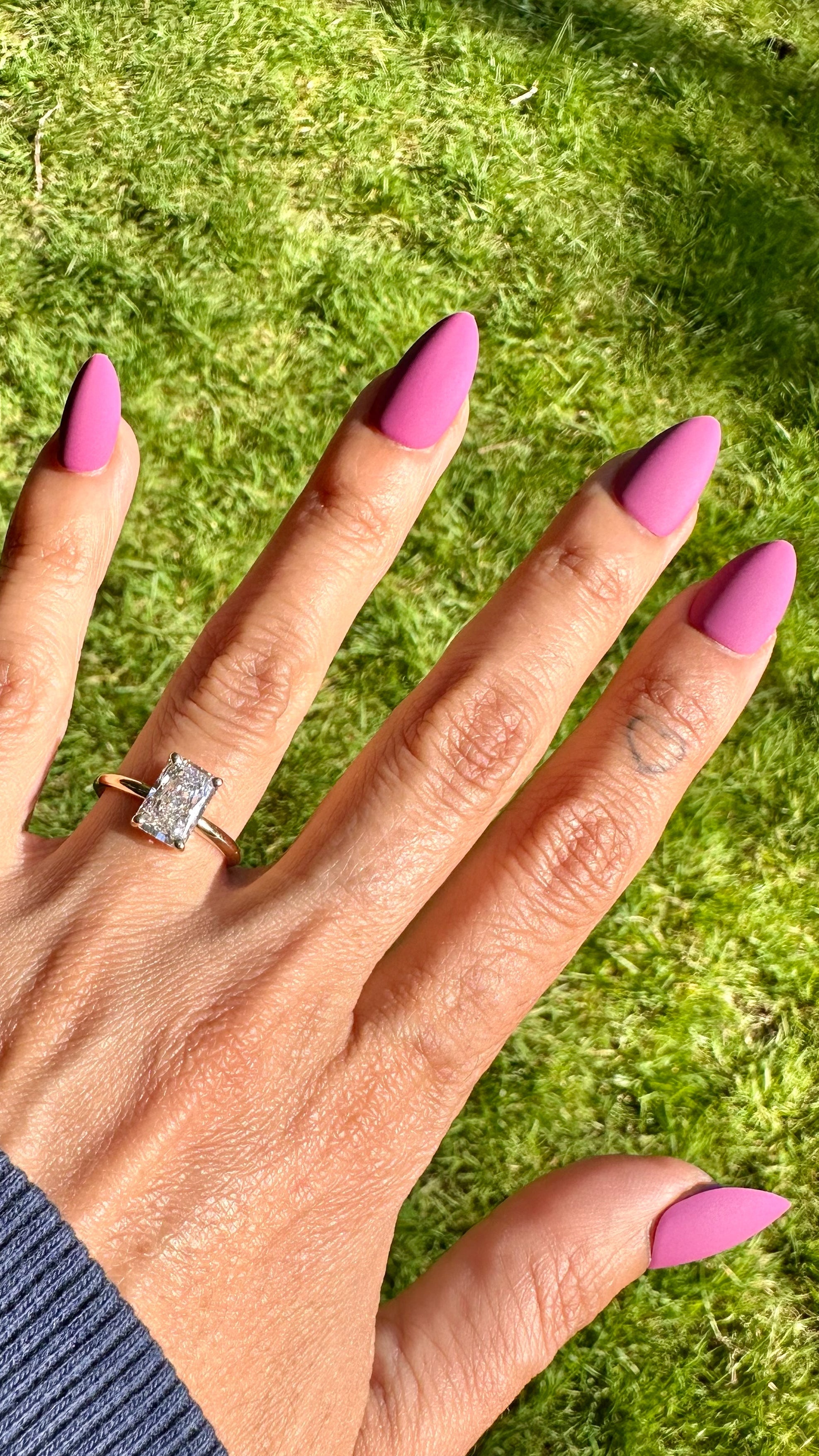 A close-up of a hand with long pink manicured nails wearing a radiant cut diamond engagement ring on a grassy lawn.