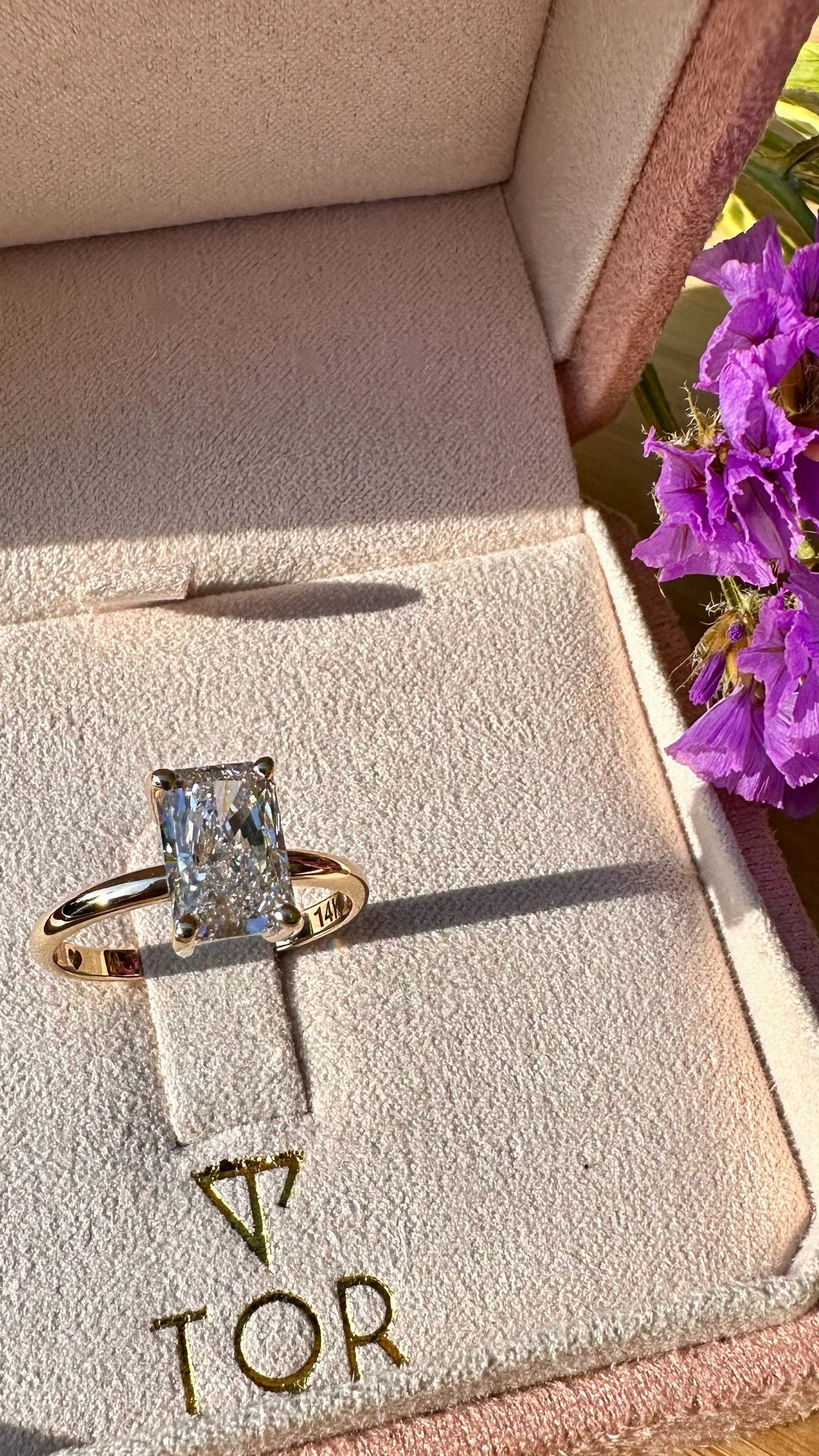 A gold ring with a radiant cut white diamond displayed inside a pink jewelry box. The box has the brand name TOR printed on the velvet lining.