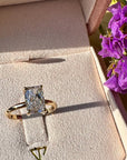 A gold ring with a radiant cut white diamond displayed inside a pink jewelry box. The box has the brand name TOR printed on the velvet lining.