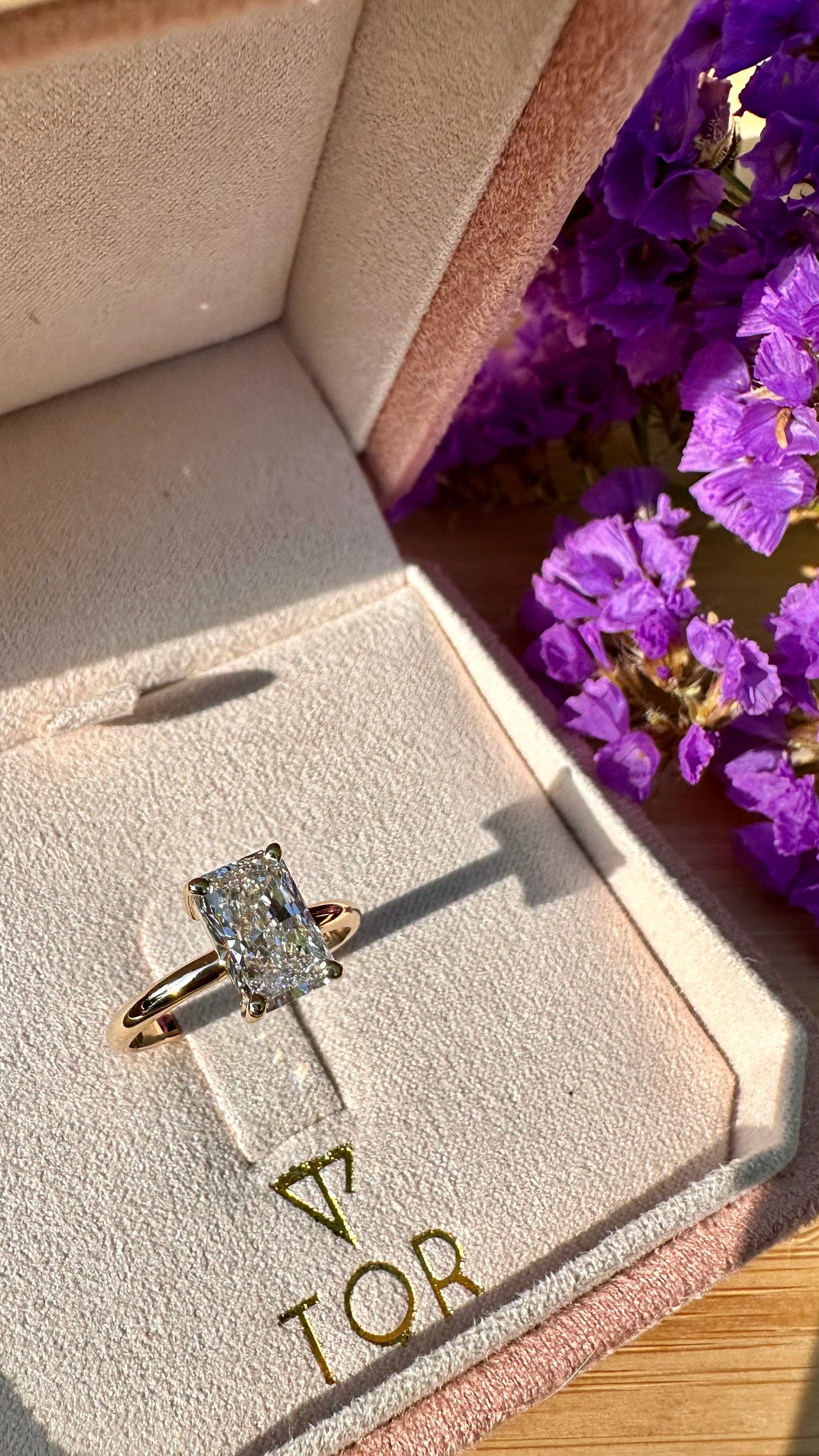 A gold ring with a radiant cut lab grown diamond displayed inside a pink velvet jewelry box with the brand name TOR printed on the inside lid. Purple flowers are visible on the right side.